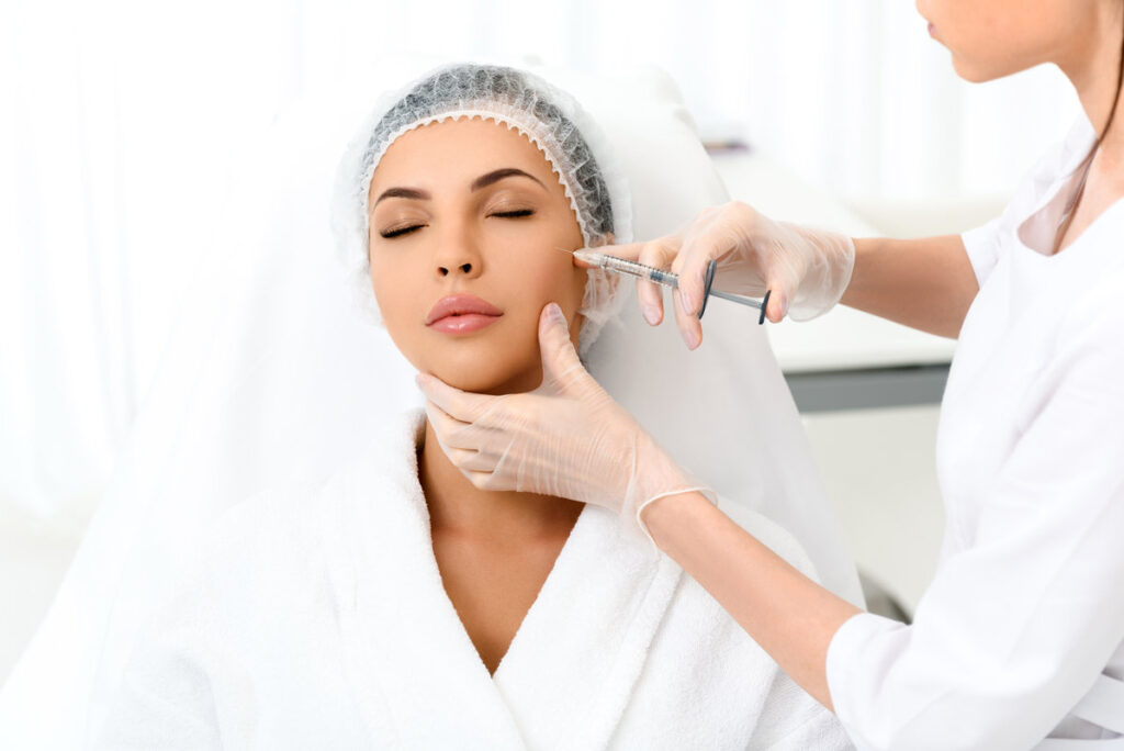 Relaxed young woman is getting botox injection at clinic. She is sitting and closed eyes. Doctor is standing and touching syringe needle to her cheek